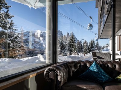 Hotel Schmung – Ihr Hotel auf der Seiser Alm Blick aus einem Fenster auf schneebedeckte Berge und Gondeln mit Sofa im Vordergrund