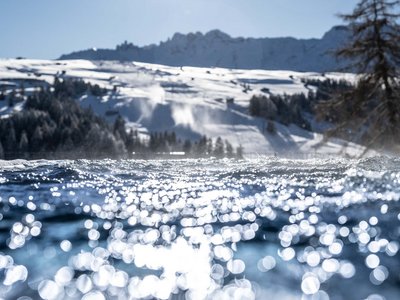 Hotel Schmung – Ihr Hotel auf der Seiser Alm Glitzerndes Wasser im Vordergrund vor verschneiten Bergen und Bäumen