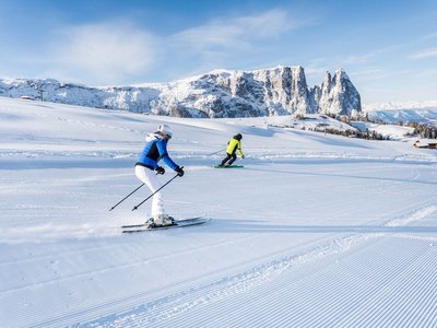 Hotel Schmung – Ihr Hotel auf der Seiser Alm Zwei Skifahrer fahren auf einer präparierten Piste vor schneebedeckten Bergen