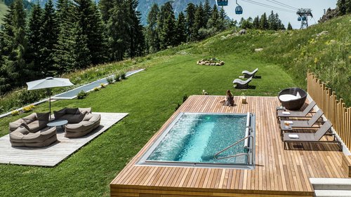 Hotel Schmung – Ihr Hotel auf der Seiser Alm Poolbereich mit Liegestühlen und Bergseilbahn auf einer grünen Wiese im Sommer