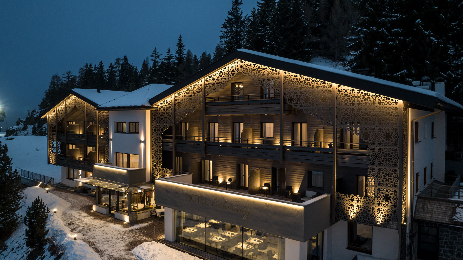 Hotel Schmung – Ihr Hotel auf der Seiser Alm Beleuchtetes Alpenhotel im Schnee, umgeben von dunklen Tannen bei Dämmerung