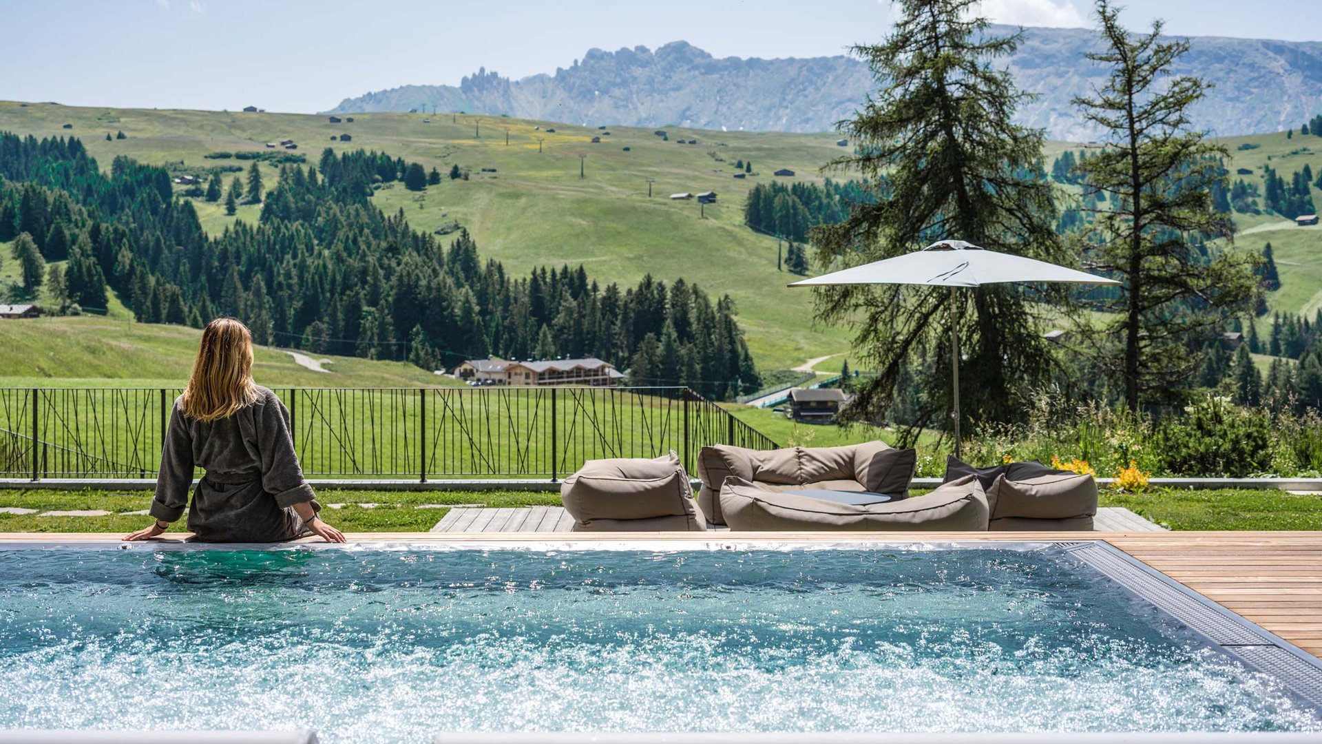 Hotel Schmung – Ihr Hotel auf der Seiser Alm Frau entspannt am Pool mit Bergpanorama und Sitzbereich im Grünen
